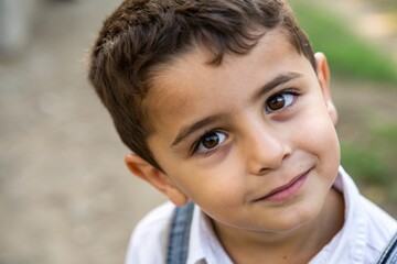 Young boy portrait, innocent smile, big brown eyes, close-up, natural lighting, soft focus, outdoor setting, blurred background, white shirt, candid expression, warm tones, Mediterranean features