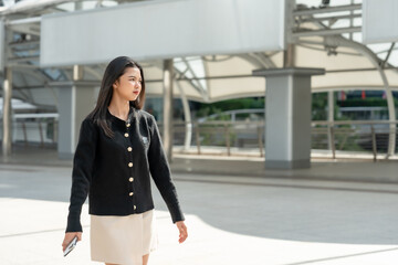 Beautiful Asian office worker walking in business district. Young woman in elegant black blazer walking confidently in modern business area holding smartphone in professional urban setting.