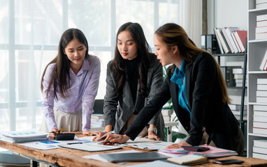 Professional women collaborating, reviewing financial graphs near wooden table, modern office interior with natural light and bookcase backdrop
