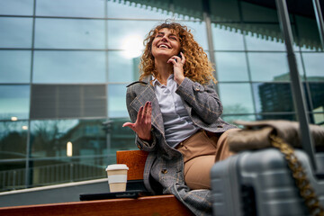 Sitting on a bench, a Caucasian woman uses her phone, holding a takeaway coffee with a suitcase...