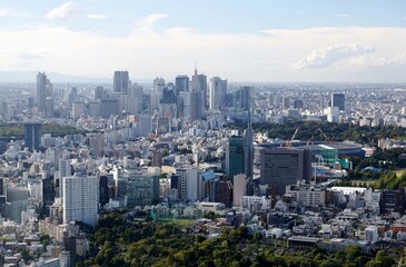 Aerial panorama of Downtown Tokyo, with a city skyline of high-rise office towers clustering in Shinjuku District and an urban park among crowded buildings under sunny cloudy sky