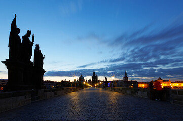 Scenery of Charles Bridge before sunrise with street lamp lights glowing in blue twilight & silhouettes of majestic statues & towers of historical buildings under dawning sky in Old Town Prague, Czech