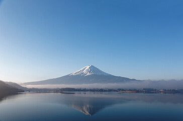 Scenery of snow capped Mount Fuji reflected in the peaceful water of Kawaguchi Lake with dense fog lingering at the foothill under blue clear sky on a sunny winter morning, in Yamanashi, Japan