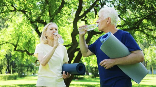 Elderly man and woman drink water from bottles while holding yoga mats in green park. Refreshment after physical activity - Powered by Adobe
