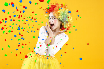 Happy girl dressed like clown under falling confetti on orange background. Party time