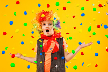 Cute boy dressed like clown under falling confetti on orange background. Party time