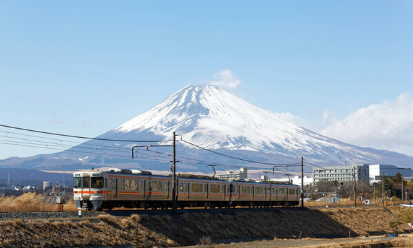 A local train of JR Gotemba Line (御殿場線) traveling through the countryside on a sunny winter day and snow capped Mount Fuji dominating the background under blue clear sky in Susono, Shizuoka, Japan