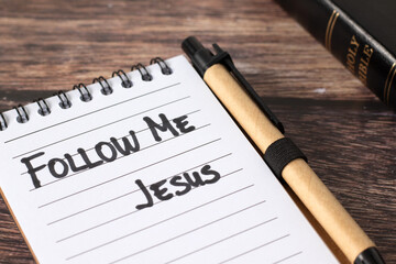 Follow Me, Jesus, handwritten quote in notebook with holy bible book on wooden background. Close-up. Christian obedience, direction, faith, and guidance, biblical concept.