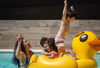Excited latin family playing on inflatable duck float in swimming pool, enjoying summer vacation