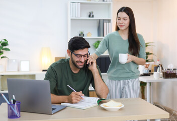 Image of a handsome white man looking at a laptop and working online from home with his girlfriend making him coffee.