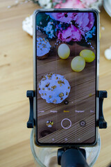 A photographer shoots an Easter composition on a phone. In the frame, Easter cakes, Easter eggs, flowers on a wooden table, a camera on a tripod.