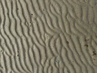 Close-up view of sand with wavy patterns.
The sand is a light gray or beige color, and the texture is fine-grained. The wavy patterns are created by the movement of the sand, likely by wind or water.