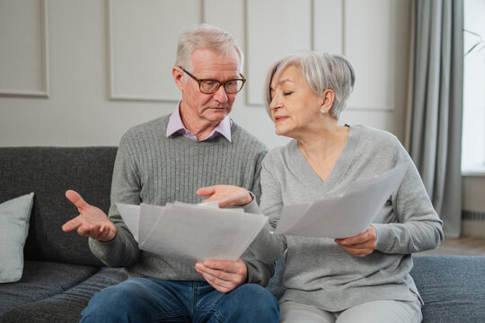 Sad tired disappointed middle aged senior couple sit with paper document. Unhappy older mature man woman reading paper bill managing bank finances calculating taxes planning loan debt pension payment