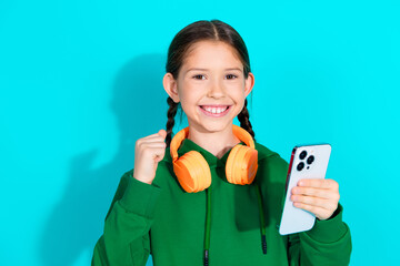 Charming young girl with headphones holding a smartphone against a teal background, depicting joy...