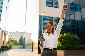 Businesswoman celebrating success outdoors holding coffee cup and smartphone