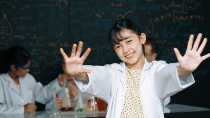 Cute asian scientist waving hand to camera while diverse group doing experiment. Young schoolgirl...