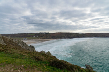Vue sur la plage de la baie des Trépassés depuis les falaises, avec la mer calme, des vagues laissant une écume blanche et un ciel cotonneux qui se mêle à l'horizon.