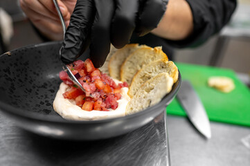 A chef's hand with black gloves carefully arranges diced ingredients on a dish. The setting features a stainless steel surface and chopping board, highlighting culinary creativity.