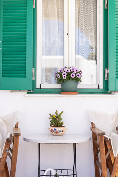 Charming vignette of a Greek patio with green windows, purple flowers, and soft chairs. 