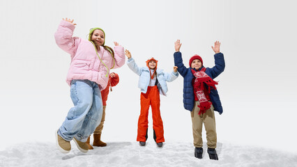 Obraz premium Group of children, girls and boys, dressed warm winter clothes running, holding hands while smiling and enjoying winter fun against white studio background. Concept of winter vacation, friendship.