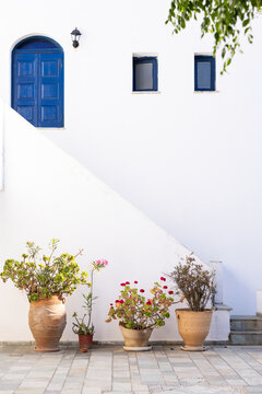 Picturesque facade of a Greek home with blue door and windows, and terra cotta pots of flowers against a whitewashed wall. 