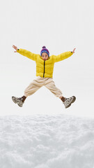 Boy in yellow puffer jacket, beige pants, blue hat jumping high with stretched arms, playful...