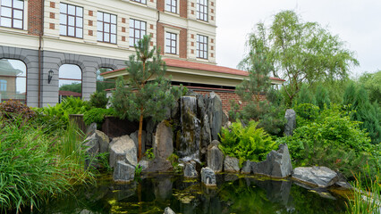 Green courtyard, pond, rocks and waterfall on embankment of Iset River, Yekaterinburg, Russia. Urban landscaping in residential area. Garden next to rickety house