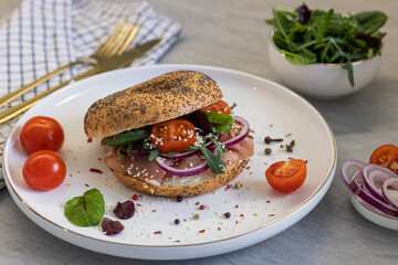 Tasty poppy seed bagel with smoked meat, creamy cheese, cherry tomatoes, red onion, and fresh arugula on a white plate with spices. Healthy gourmet breakfast, homemade meal and fresh ingredients