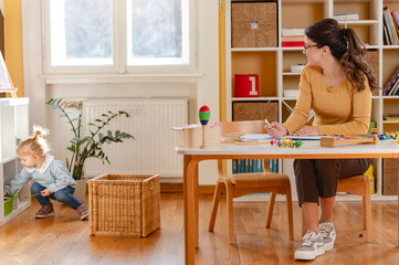 A preschool teacher writes notes at a desk while a curious toddler girl explores a shelf in a...