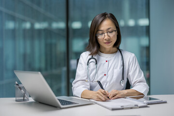 Serious thinking female doctor working with papers and documents inside clinic office. Asian woman in white medical coat and with stethoscope recording patient data. Woman preparing treatment report.