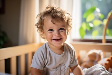 A cheerful toddler with curly hair sits in a wooden crib, beaming with joy. Soft sunlight filters through the window, illuminating the cozy room filled with plush toys and green plants.