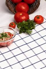 ensalada chilena, tomate cebolla y perejil, junto a tomates maduros y ramas de perejil, sobre una tela blanca con lineas azules, vista cenital