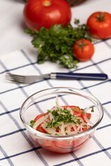 ensalada chilena, tomate cebolla y perejil, al fondo un tenedor, tomates maduros y ramas de perejil sobre un paño de cocina blanco con lineas azules