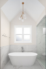 A bathroom freestanding soaking tub in a luxury bathroom with a gold chandelier hanging from the tall ceiling, tan walls, marble tiled flooring, and a white marble tiled shower.