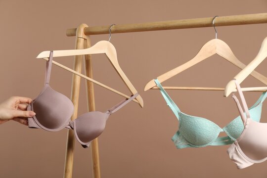 Woman choosing beautiful bra near rack on dark beige background, closeup