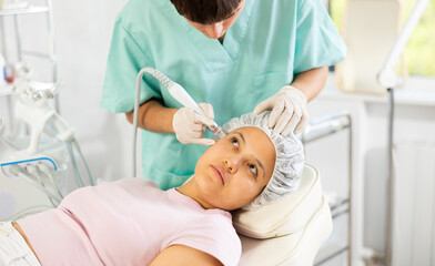 Young male cosmetologist performs hardware facial cleansing procedure to young woman patient