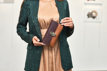 Elegant woman holding stylish leather clutch in green jacket and beige dress, showcasing fashion accessory elegance concept