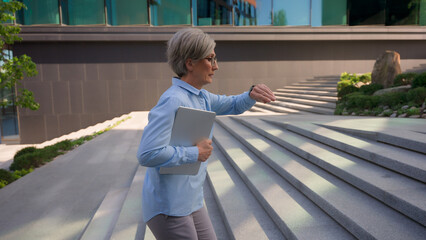 Caucasian mature business woman hurries walking up stairs office building in city looking at hand...