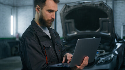 Serious focused Caucasian man guy technician auto mechanic engineer check repairing car problem computer diagnostics electric automobile engine with laptop checking maintenance in garage vehicle shop