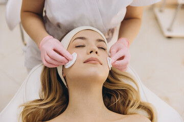 Cosmetologist wearing pink gloves is cleansing skin of client with cotton pads during facial treatment in spa