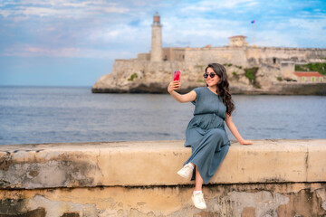 Fototapeta premium Young Latina woman in a blue dress and sunglasses takes a selfie with a red smartphone while sitting on a stone wall near Morro Castle in Havana, Cuba. 