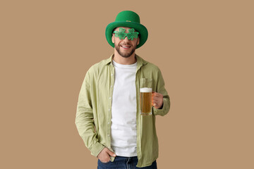 Young man in leprechaun hat and decorative glasses in shape of clover with glass of beer on brown...