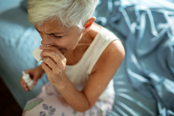 Senior woman sneezing into tissue while sitting on bed with cold or allergies