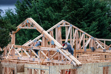Workman on a ladder on second floor of new home under construction nailing a new wood roof truss...