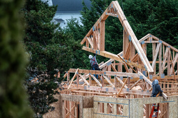 Workman on a ladder on second floor of new home under construction guiding a wood roof truss being...