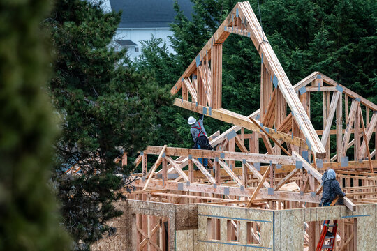 Workman on a ladder on second floor of new home under construction guiding a wood roof truss being lifted by crane into place
