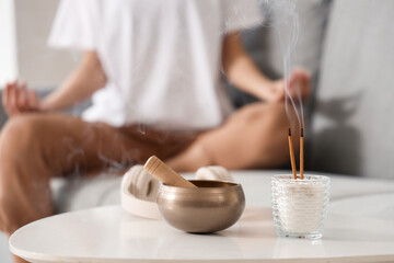 Tibetan singing bowl with aroma sticks and headphones on table against woman meditating at home, closeup