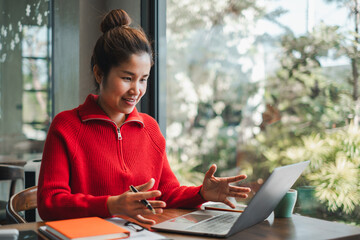 Woman in red sweater having a video call on a laptop, sitting by a window with greenery outside.