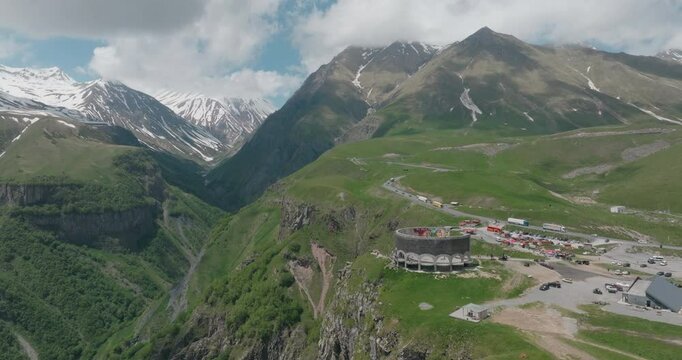 Drone shot passing through the Georgia-Russia Friendship Monument. Surrounded by the peaks of the Caucasus Mountains and lush green valleys, this historical building offers an impressive view.