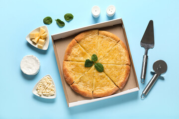 Cardboard box with tasty pizza, cheese, basil leaves, knife and spatula on blue background